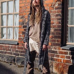 May include: A person wearing a gray hooded cardigan, a brown shirt, and brown pants. The cardigan has a black and white pattern. The person is also wearing a gray beanie and black shoes with red laces. The background is a brick wall.