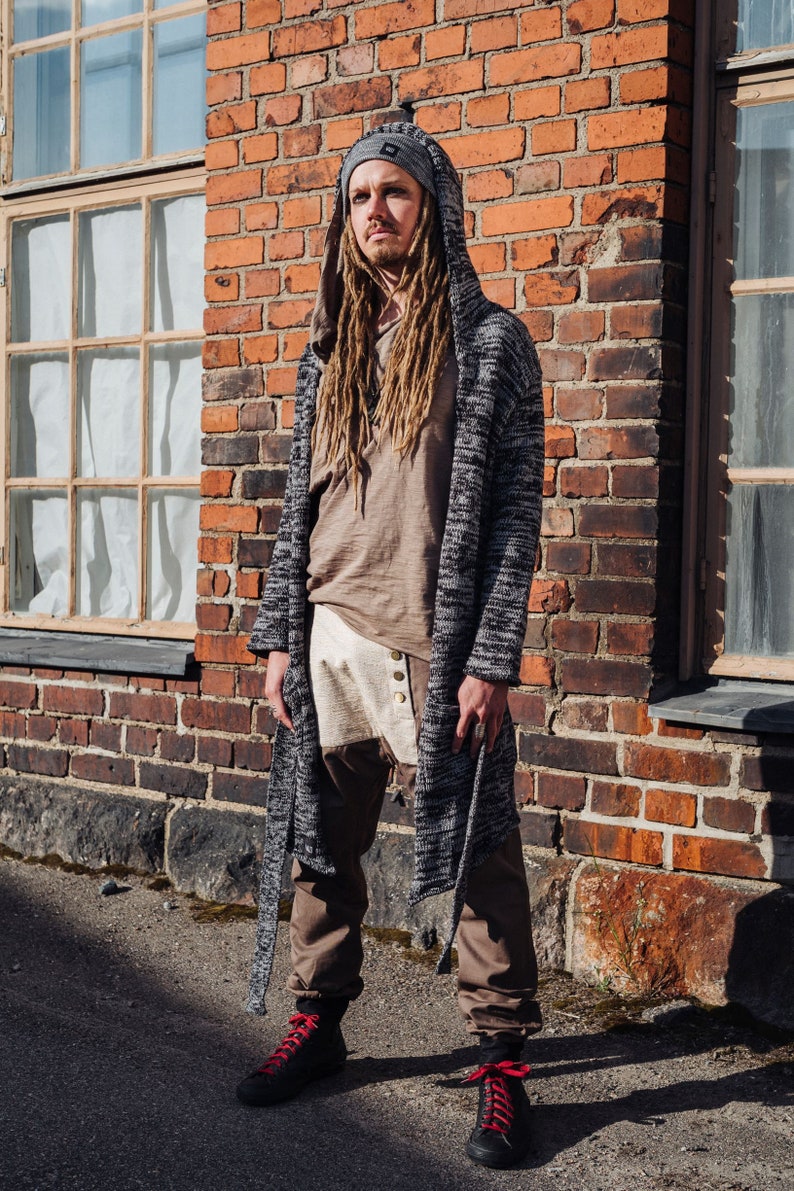 May include: A person wearing a gray hooded cardigan, a brown shirt, and brown pants. The cardigan has a black and white pattern. The person is also wearing a gray beanie and black shoes with red laces. The background is a brick wall.