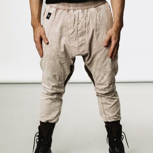 May include: Light beige drop-crotch pants with a subtle pattern, featuring a drawstring waist and knee paneling. The pants are paired with black combat boots. The model is wearing a black t-shirt.