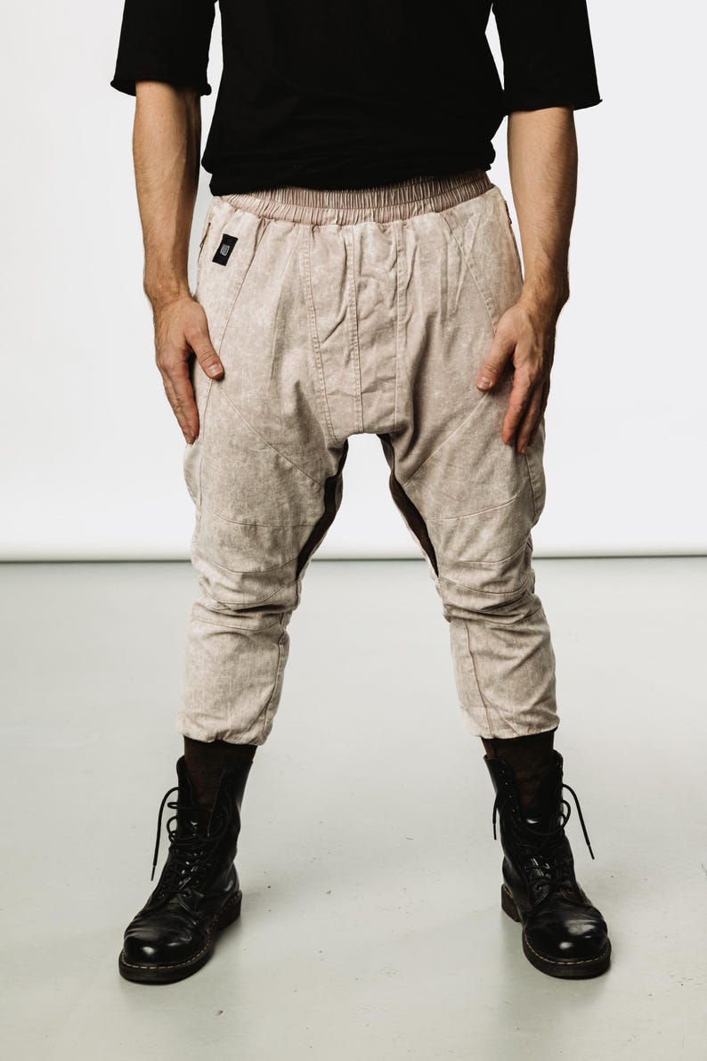 May include: Light beige drop-crotch pants with a subtle pattern, featuring a drawstring waist and knee paneling. The pants are paired with black combat boots. The model is wearing a black t-shirt.