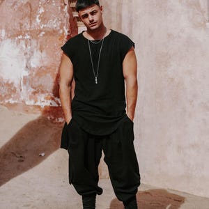 May include: A person wearing a black sleeveless top, black pants, and black high-top sneakers. The person is also wearing a silver necklace with a pendant. The background is a textured, light-colored wall.