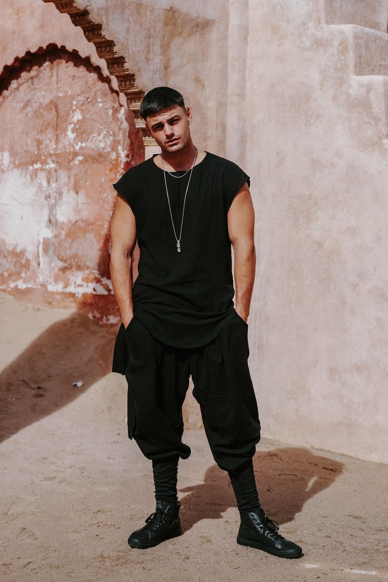 May include: A person wearing a black sleeveless top, black pants, and black high-top sneakers. The person is also wearing a silver necklace with a pendant. The background is a textured, light-colored wall.