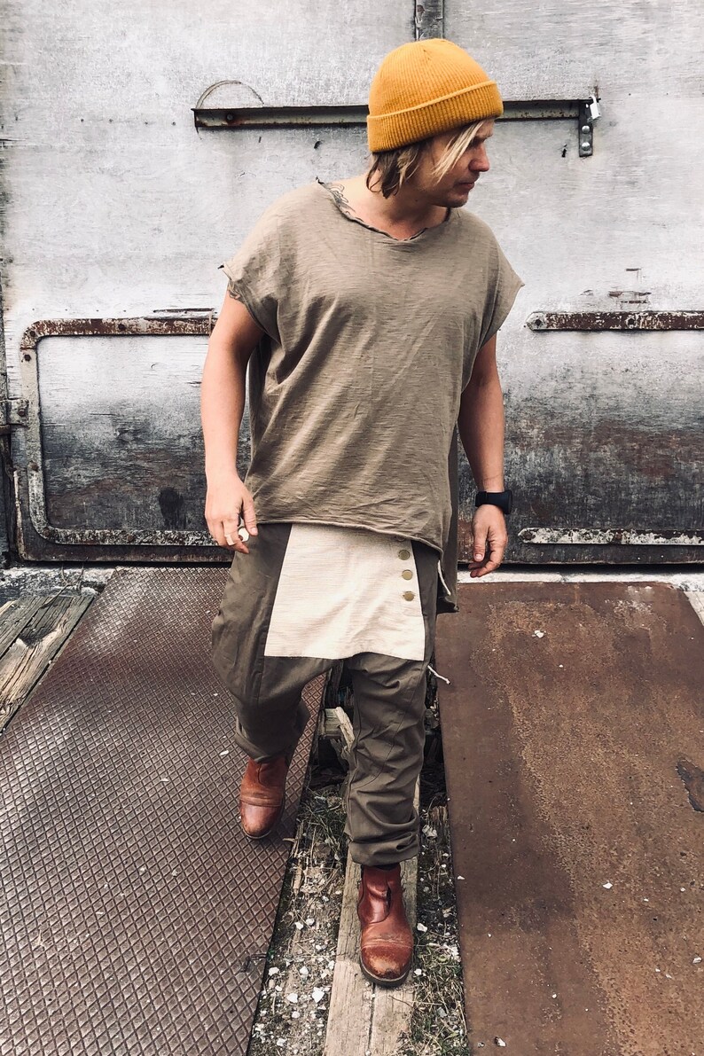 May include: A person wearing a mustard yellow beanie, a loose-fitting olive green t-shirt, and olive green pants with a cream-colored panel. The person is also wearing brown leather boots. The outfit has a casual, bohemian style.