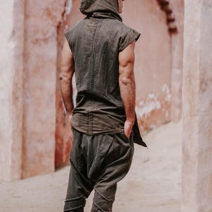 May include: A dark olive green hooded tunic and matching harem pants. The sleeveless tunic has a hood and a long back panel. The pants are gathered at the ankles. The outfit is made of a soft, flowing fabric. The person is standing in front of a pinkish-red wall.