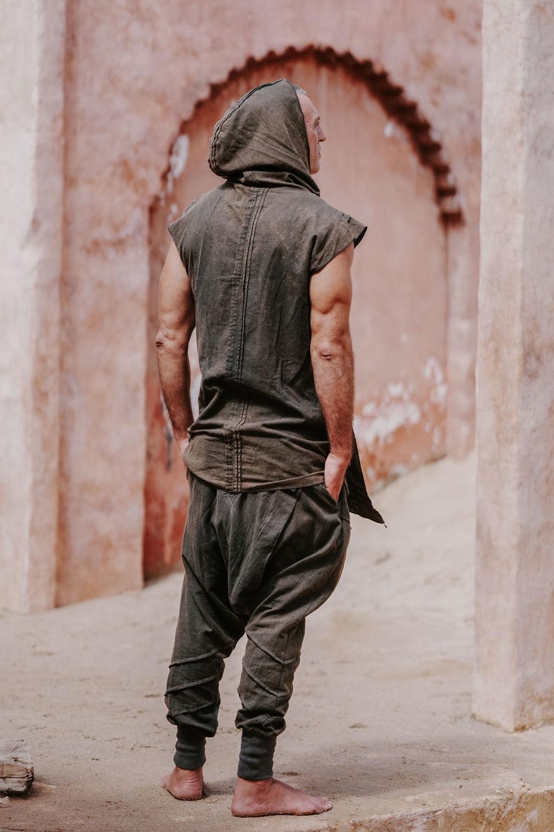 May include: A dark olive green hooded tunic and matching harem pants. The sleeveless tunic has a hood and a long back panel. The pants are gathered at the ankles. The outfit is made of a soft, flowing fabric. The person is standing in front of a pinkish-red wall.
