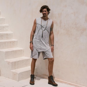 May include: A man wearing a grey sleeveless shirt with a long beaded necklace, grey shorts, and brown boots. He is standing in front of a white wall with stairs.