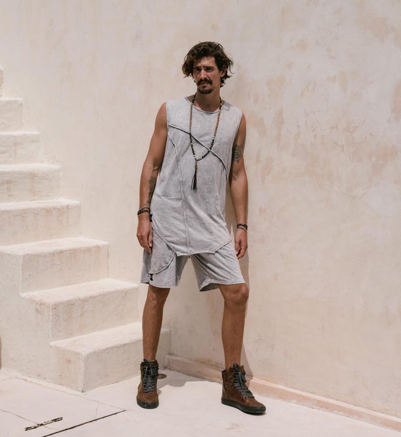 May include: A man wearing a grey sleeveless shirt with a long beaded necklace, grey shorts, and brown boots. He is standing in front of a white wall with stairs.