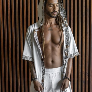 May include: A white hooded robe with brown and white patterned trim. The robe is open in the front, revealing a white undershirt and a tattoo on the chest that reads "Elevate".