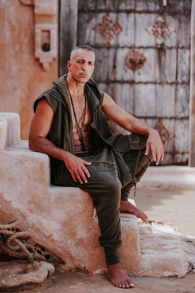 May include: A man wearing a sleeveless olive green hooded vest, matching pants, and a beaded necklace. He is seated on a stone structure with a weathered wooden door in the background. The outfit has a relaxed, bohemian style.