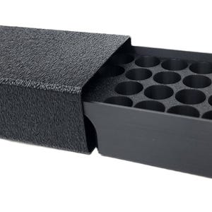 May include: A black rectangular storage case with a textured exterior finish and a sliding drawer. The drawer features a grid of twenty-four circular compartments for organizing small items. A small silver-colored metal knob is on the side.