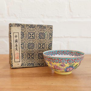 May include: A decorative porcelain bowl with a yellow base and floral design, alongside a patterned box. The bowl has a turquoise rim and a floral pattern with pink and blue flowers. The box has Chinese text.