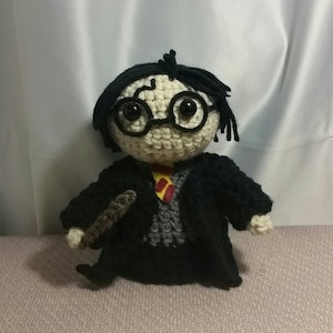 Puede incluir: Una muñeca de ganchillo con cara pálida, pelo de lana negra, gafas redondas negras y una cicatriz en forma de rayo en la frente. La figura lleva una túnica negra sobre un suéter gris y una corbata a rayas amarillas y rojas, sosteniendo una varita marrón.