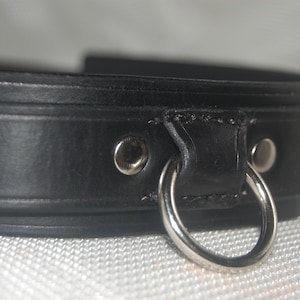 Pode incluir: Close-up de uma coleira de couro preta com um anel e rebites de metal prateado. A coleira &eacute; feita de couro grosso e liso e parece ser bem trabalhada. O anel de metal &eacute; preso com costuras resistentes.