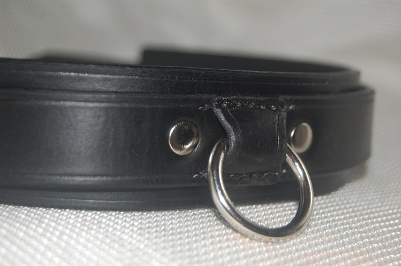 Pode incluir: Close-up de uma coleira de couro preta com um anel e rebites de metal prateado. A coleira &eacute; feita de couro grosso e liso e parece ser bem trabalhada. O anel de metal &eacute; preso com costuras resistentes.