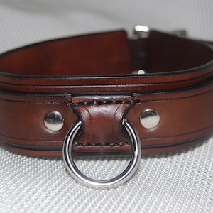 Pode incluir: Uma coleira de couro castanho com um anel e fivela de metal prateado. A coleira &eacute; espessa e tem um design detalhado com rebites prateados. O couro tem uma cor castanha escura rica.