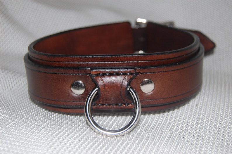 Pode incluir: Uma coleira de couro castanho com um anel e fivela de metal prateado. A coleira &eacute; espessa e tem um design detalhado com rebites prateados. O couro tem uma cor castanha escura rica.
