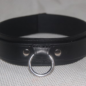 Pode incluir: Uma coleira de couro preta com um anel de metal prateado e dois rebites prateados. A coleira &eacute; grossa e tem uma borda arredondada. O fundo &eacute; uma superf&iacute;cie texturizada cinza claro.