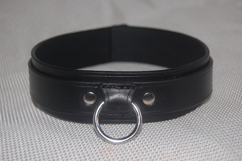 Pode incluir: Uma coleira de couro preta com um anel de metal prateado e dois rebites prateados. A coleira &eacute; grossa e tem uma borda arredondada. O fundo &eacute; uma superf&iacute;cie texturizada cinza claro.