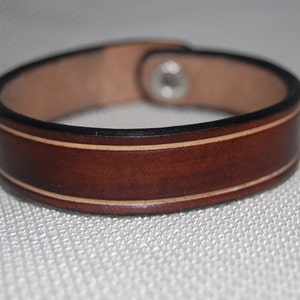 Könnte beinhalten: Ein braunes Lederarmband mit einem weißen, gesteppten Rand. Das Armband hat einen Metallverschluss.