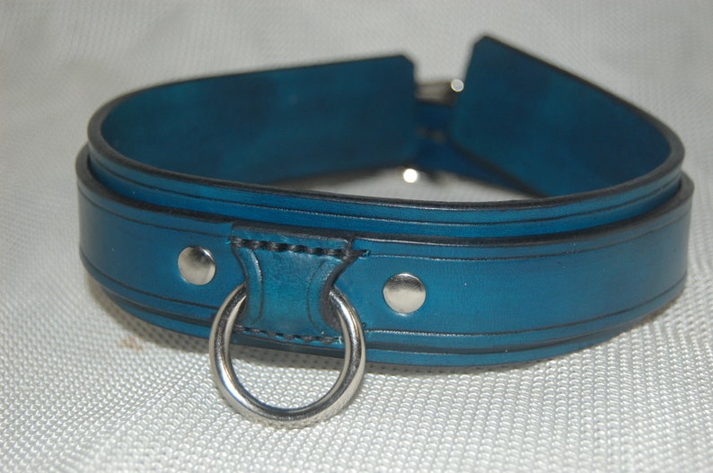 Pode incluir: Uma coleira de couro azul-petr&oacute;leo com um anel e rebites de metal prateado. A coleira tem um design arredondado com uma borda preta. O anel de metal &eacute; preso a uma se&ccedil;&atilde;o refor&ccedil;ada.