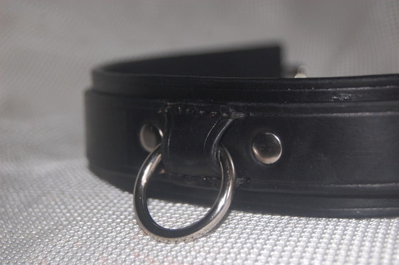 Pode incluir: Close-up de uma coleira de couro preta com um anel e rebites de metal prateado. A coleira &eacute; grossa e parece bem feita, com costuras vis&iacute;veis. O anel de metal &eacute; preso &agrave; coleira com uma pequena presilha de couro.