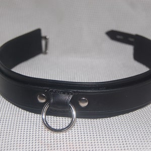 Pode incluir: Uma coleira de couro preta com um anel de metal prateado e fecho de fivela. A coleira &eacute; feita de couro preto liso e tem um design simples. O anel de metal &eacute; fixado com dois rebites prateados.