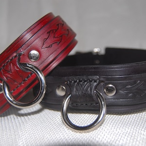 Könnte beinhalten: Zwei Lederhalsbänder mit silbernen Metallringen. Ein Halsband ist rot mit einem Blumenmuster und das andere ist schwarz mit einem Blattdesign.