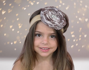May include: A young girl with long brown hair is wearing a headband with a large brown and white flower. The flower has a sparkly rhinestone in the center. The headband is a light brown color.