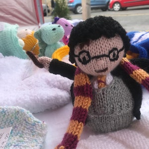 May include: Knitted Harry Potter doll with black hair, glasses, a grey sweater, and a burgundy and gold scarf. The doll is holding a wand in its right hand.