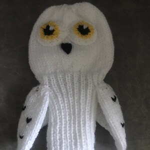 May include: White knitted owl with black eyes and yellow accents. The owl has small black hearts on its wings.