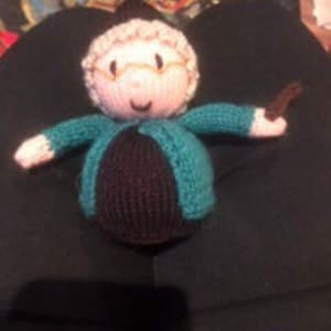 May include: A crocheted plush toy of a woman wearing a green dress, brown cardigan, and glasses. She has a brown hair bun and is holding a brown stick.