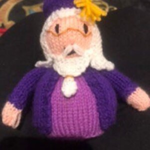 May include: A knitted purple and white doll with a yellow hat and a white beard. The doll is wearing a purple sweater and a white shirt.