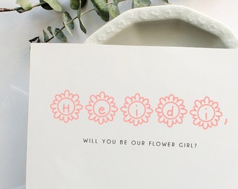 May include: A white invitation card with the name "Heidi" printed in pink floral letters. The text "Will you be our flower girl?" is below the name. An envelope and eucalyptus sprig are also present.
