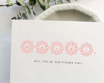 May include: A white invitation card with the name "Heidi" printed in pink floral letters. The text "Will you be our flower girl?" is below the name. An envelope and eucalyptus sprig are also present.