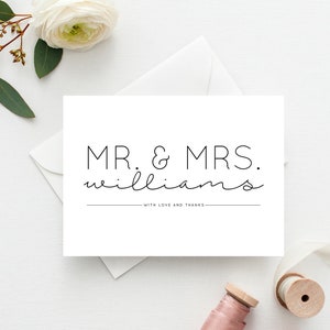 Könnte beinhalten: Eine weiße Karte mit dem Text "MR. & MRS. WILLIAMS" in einer minimalistischen Schriftart. Unter den Namen steht der Satz "WITH LOVE AND THANKS". Die Karte ist von einem weißen Umschlag, einer weißen Blume und einer Garnrolle umgeben.