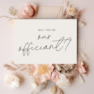 May include: A rectangular cream-coloured card featuring the black printed text "WILL YOU BE our officiant?" centred on the front. The card is surrounded by a decorative arrangement of soft pink, peach, and white flowers and dried foliage.