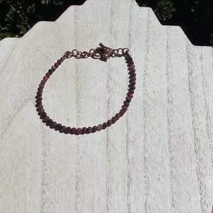 May include: A delicate bracelet featuring small, round, dark red beads strung together. The bracelet has a copper-colored clasp and chain. The jewelry is displayed on a light-colored wooden surface.