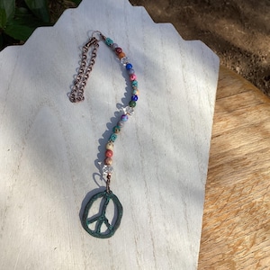 May include: A beaded peace sign charm with a copper chain and a variety of colorful beads. The peace sign is a dark teal color. The beads are in shades of blue, pink, orange, and clear.