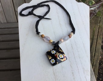 May include: A black necklace with a square pendant featuring white flowers and gold accents. The necklace is made with a black cord and has natural stone beads in shades of brown, pink, and gray.