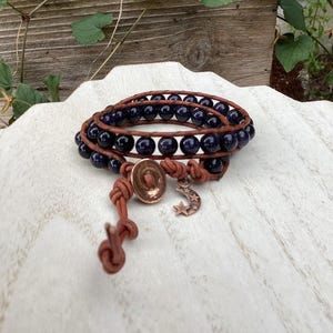 May include: A multi-wrap bracelet featuring dark blue spherical beads strung on brown leather cord. The bracelet includes a round copper-toned button closure and a small dangling charm shaped like a crescent moon with three small stars.