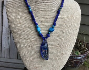 May include: A blue and turquoise necklace with a glass jellyfish pendant. The necklace is made with blue cord and turquoise beads.