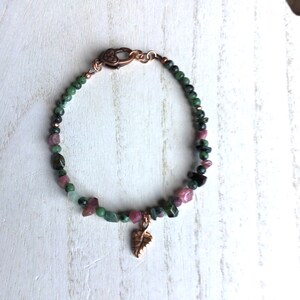May include: A green and pink gemstone bracelet with a copper leaf charm. The bracelet is made with small, irregular shaped stones and has a copper clasp.