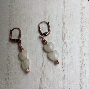 May include: A pair of handmade earrings featuring two heart-shaped, off-white gemstones suspended from copper-colored lever-back closures. The gemstones are connected with copper wire, creating a delicate and romantic design. The earrings are displayed on a light-colored wooden surface.