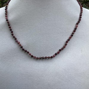 May include: A necklace made of small, round, dark red beads. The beads are strung on a thin cord and the necklace is displayed on a white background.