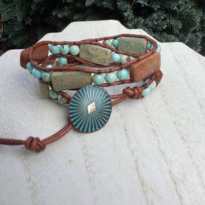 May include: A wrap bracelet featuring turquoise beads, rectangular stone accents, and brown leather straps. The bracelet has a turquoise and silver button closure. The jewelry is displayed on a white wooden surface.