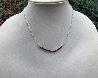 May include: A silver necklace with a delicate chain and a curved bar of small, dark red beads. The necklace is displayed on a white mannequin, showcasing the jewelry's design and color.