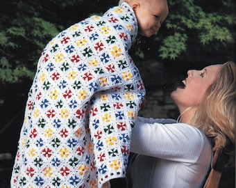 May include: A woman holding a baby wrapped in a colorful crocheted blanket. The blanket is made with white yarn and features a repeating pattern of red, blue, green, and yellow flowers.