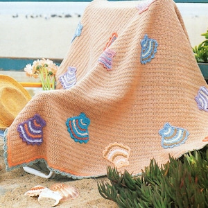 May include: A crocheted throw blanket in a light peach color with a crocheted border. The blanket has several colorful seashell designs in shades of blue, purple, orange, and green.