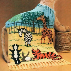 May include: A colorful crocheted tapestry featuring a giraffe, zebra, skunk, and tiger in a zoo setting. The tapestry has a light blue background with green trees and a yellow ground. The tapestry has yellow fringe around the edges.