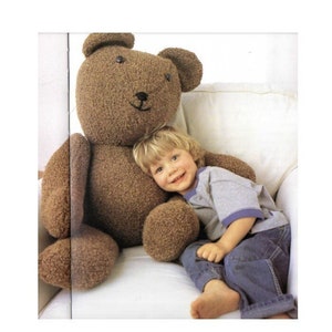 Puede incluir: Un gran oso de peluche marrón con una cara sonriente y ojos de botón. Un niño pequeño está sentado al lado del oso, apoyando la cabeza en el brazo del oso. El oso está hecho de una tela texturizada.